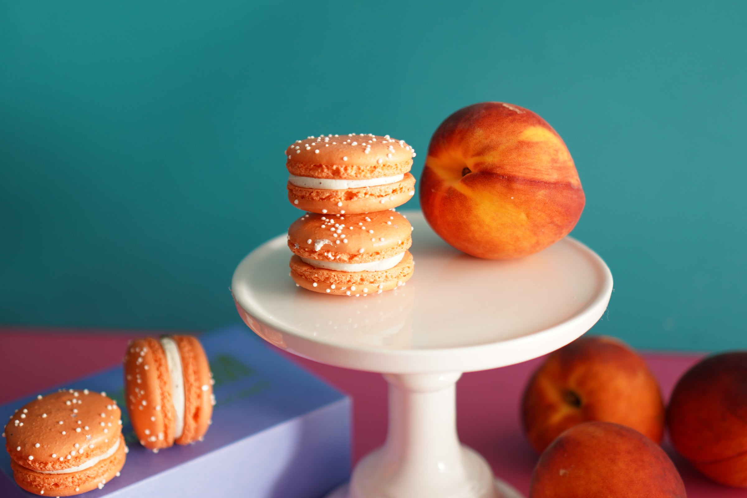 Three peach sandwich cookies with white cream and sprinkles are displayed on a white pedestal, with whole peaches and additional cookies arranged around them against a teal and pink background.