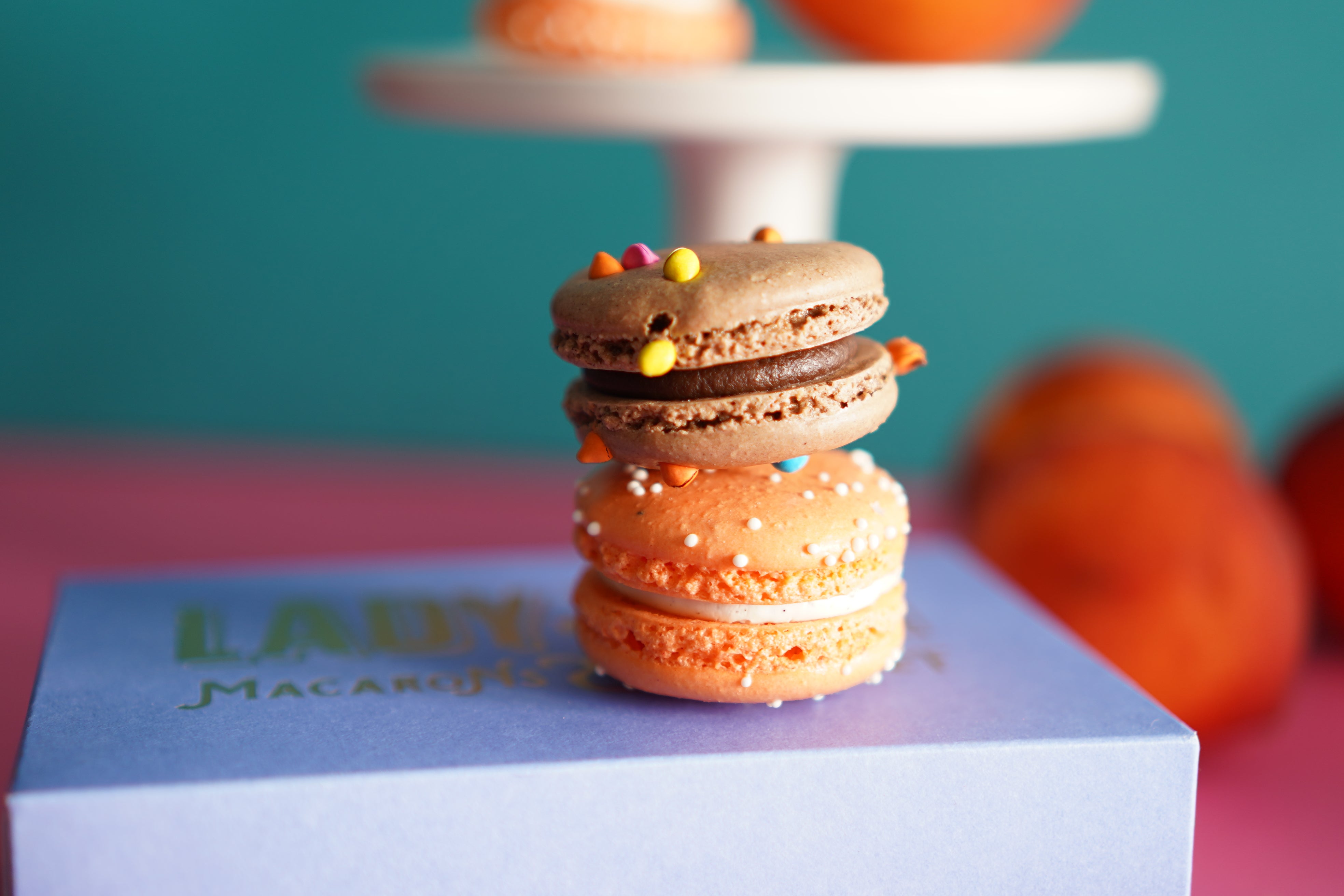 Three stacked macarons with colorful sprinkles on top, displayed on a light blue box with the text 'LADY Macarons' in green. The background is a soft-focus teal and pink setting with blurred macarons on a white stand.